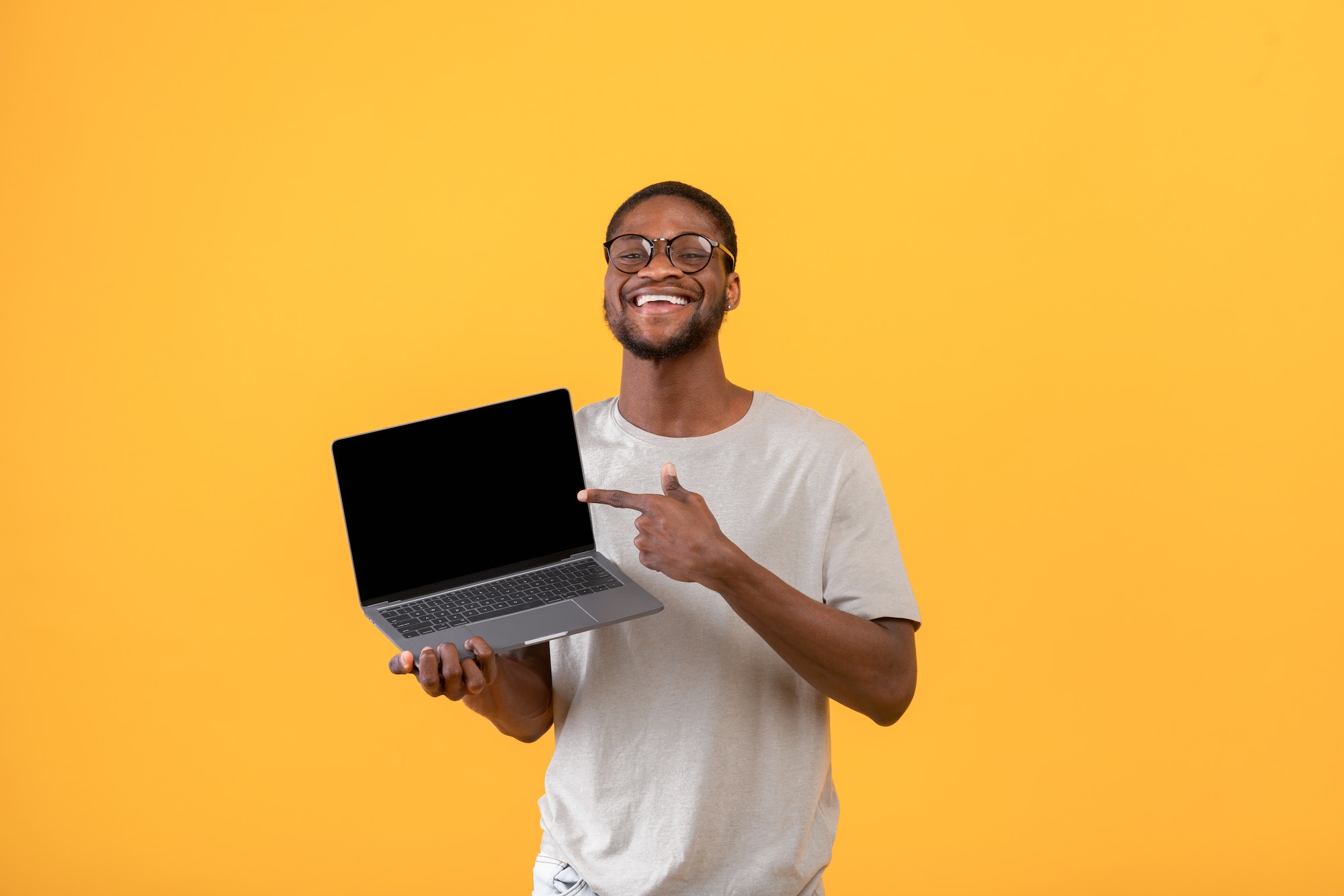 Joyful black man pointing at blank laptop screen with space for your website design over yellow
